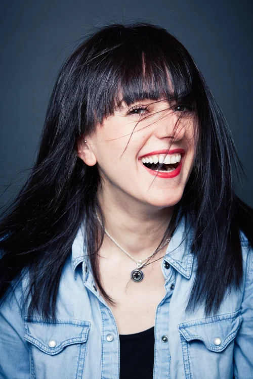A woman with long black hair and bangs, wearing a denim shirt and layered necklaces, laughing with her mouth open and eyes partially closed.