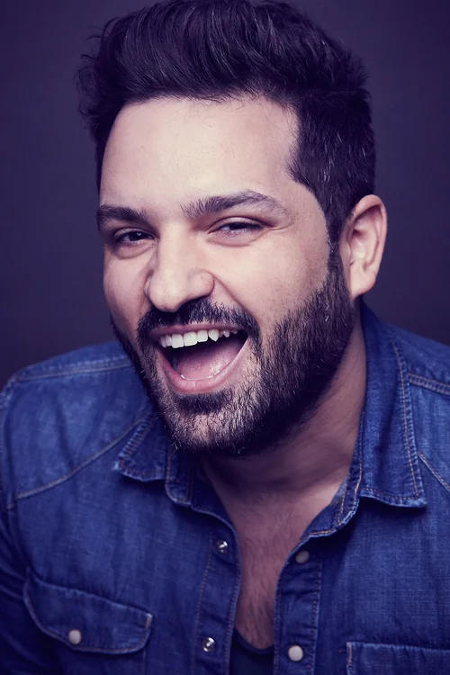 Close-up of smiling man with dark hair and beard, wearing a denim shirt, against a dark background.