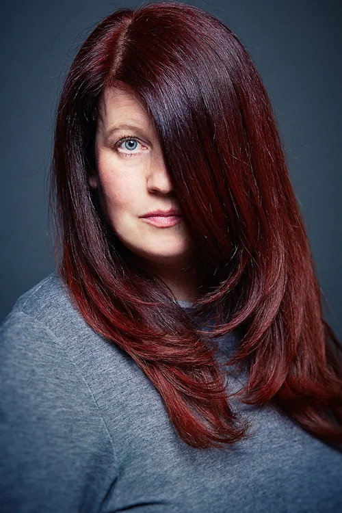 A woman with long, shiny, burgundy hair covering part of her face, looking at the camera with blue eyes and a slight smile, wearing a gray top against a dark background.