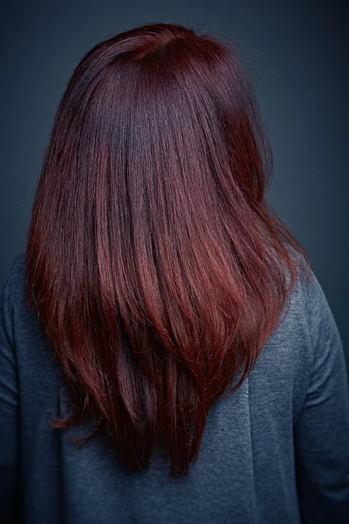 Person with straight, shoulder-length reddish-brown hair, facing downward, wearing a gray top against a dark background.