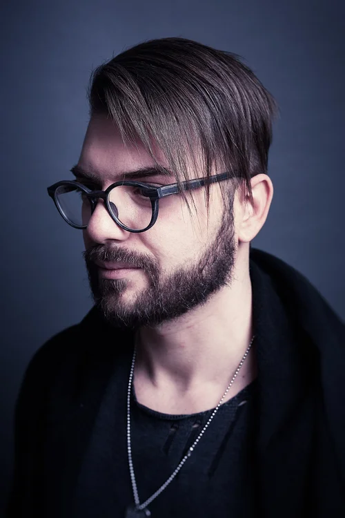 A man with  glasses and a beard, looking down, against a dark background.