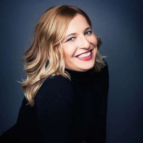 Close-up of a smiling woman with blonde, wavy hair, wearing a black top, set against a dark background.