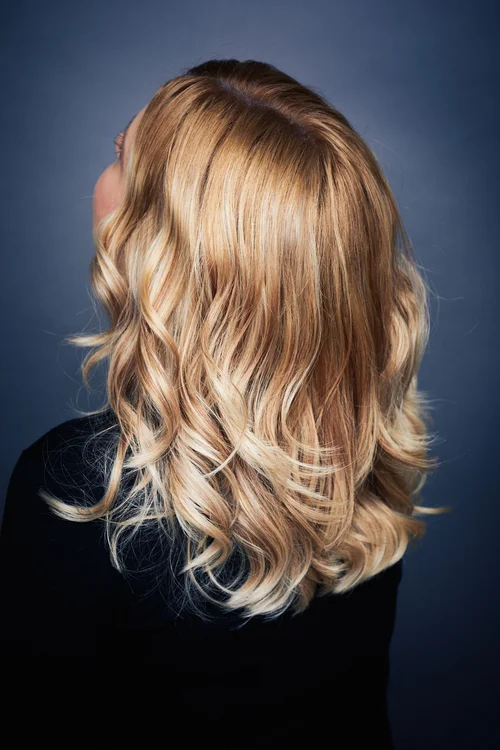 A woman with long, wavy, blonde hair standing against a dark background, wearing a black top.