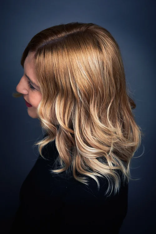 Close-up of a woman with long, wavy, blonde hair and light highlights, facing sideways against a dark background.