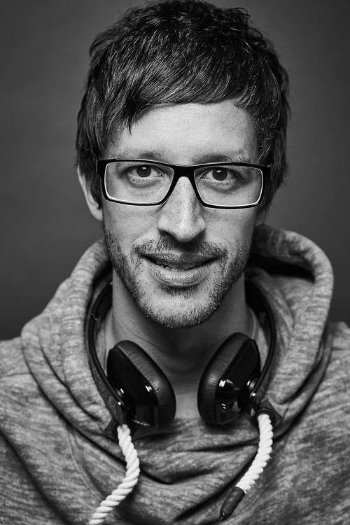 Black and white photo of a man with glasses, headphones around his neck, wearing a hoodie, smiling slightly, looking directly at the camera.
