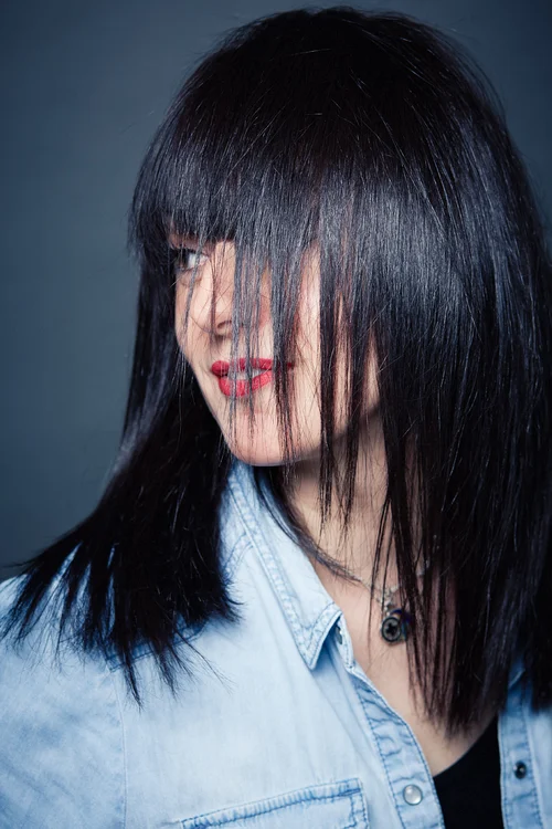 A woman with long black hair and bangs, wearing a light denim shirt and red lipstick, looking to the side against a dark background.