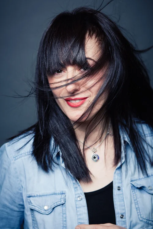 A woman with black hair and bangs, smiling with red lipstick, wearing a denim jacket and layered necklaces, with hair partially covering her face.